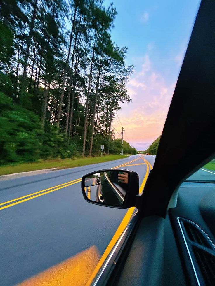 Car driving on scenic road at sunset, view from window with trees and colorful sky.