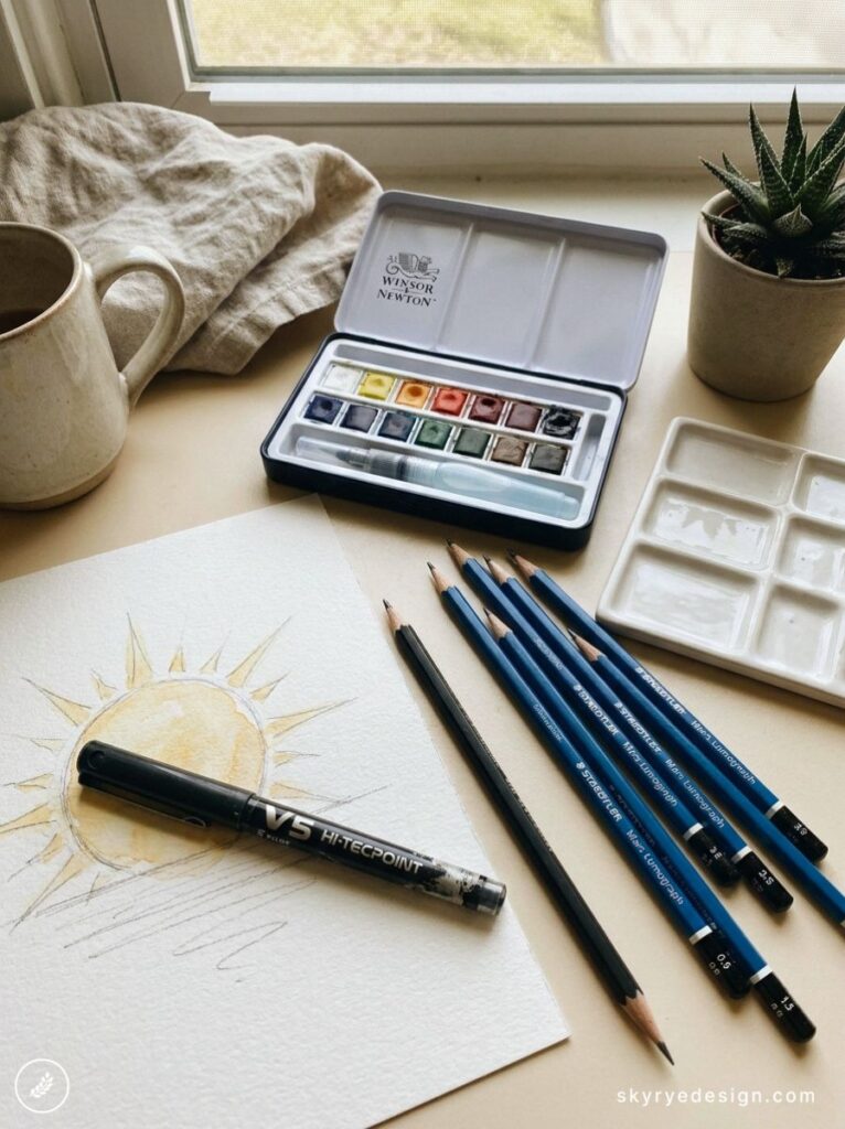Winsor & Newton watercolor set, blue drawing pencils and pen beside sun watercolor sketch on windowsill desk