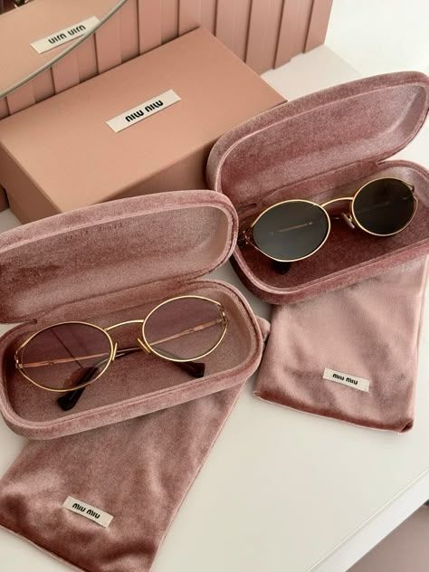 Two pairs of stylish sunglasses in pink velvet cases on a white surface, with branded packaging in the background.