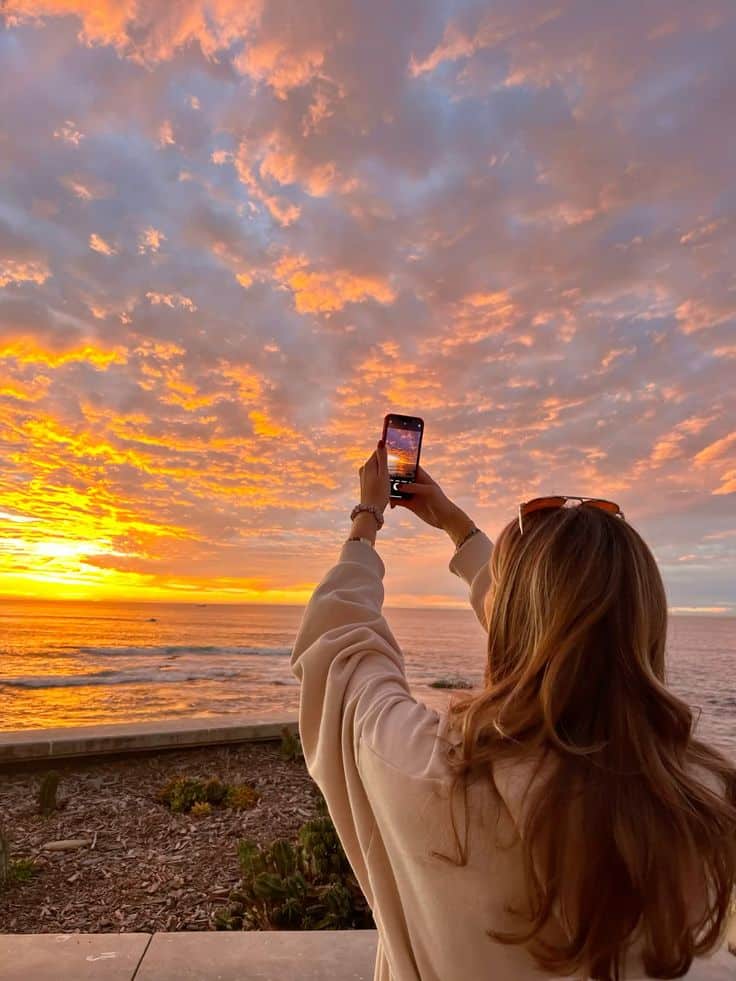 Person capturing sunset over ocean with smartphone, vibrant orange and pink sky reflecting on the water.