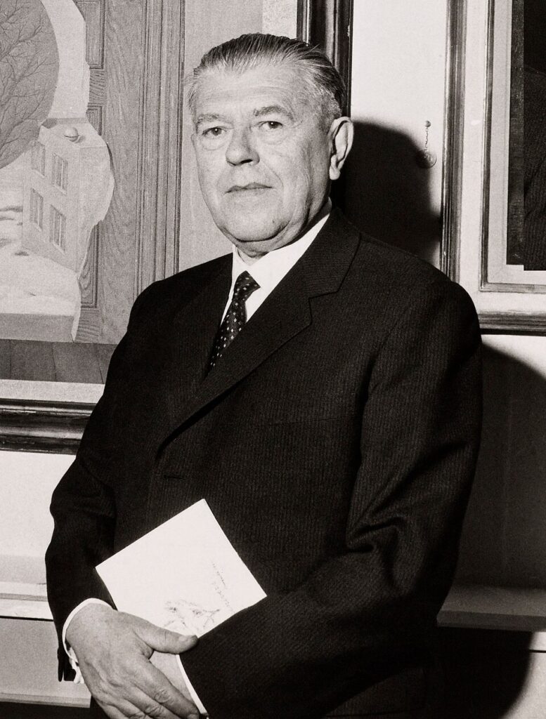 Black-and-white portrait of an older man in a suit holding a booklet, standing in front of framed artwork.