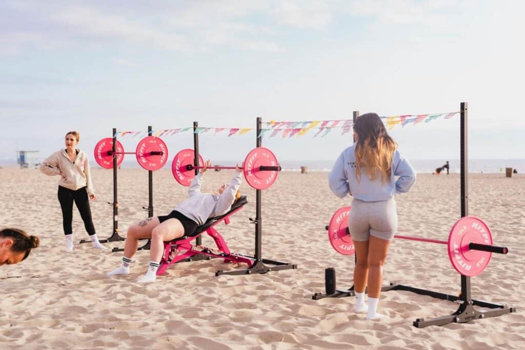 unnamed People doing beach bench press workout with pink weights under colorful flags. | Sky Rye Design People doing beach bench press workout with pink weights under colorful flags.
