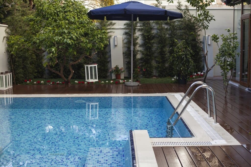 unnamed Outdoor pool with blue tiles, surrounded by lush greenery, wooden deck, and a large umbrella for shade. | Sky Rye Design Outdoor pool with blue tiles, surrounded by lush greenery, wooden deck, and a large umbrella for shade.