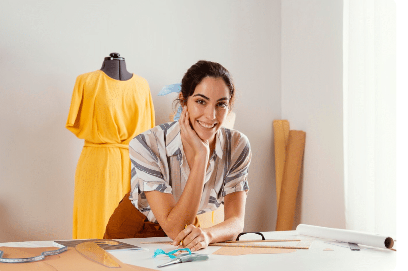 unnamed 2 Fashion designer smiling beside a dress form, surrounded by sewing tools and fabric rolls. | Sky Rye Design Fashion designer smiling beside a dress form, surrounded by sewing tools and fabric rolls.