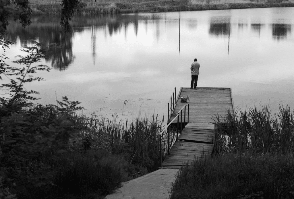 Man standing on a wooden dock fishing in a peaceful lake, surrounded by nature and reflections in the water.