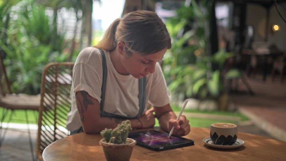 unnamed 2 Woman drawing on tablet in cafe with coffee, surrounded by plants and bright natural light. | Sky Rye Design Woman drawing on tablet in cafe with coffee, surrounded by plants and bright natural light.
