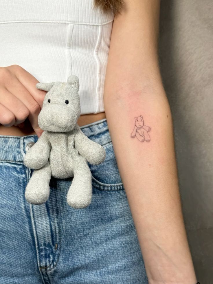 Stuffed animal tattoo on arm with matching plush toy in hand, wearing a white top and jeans.