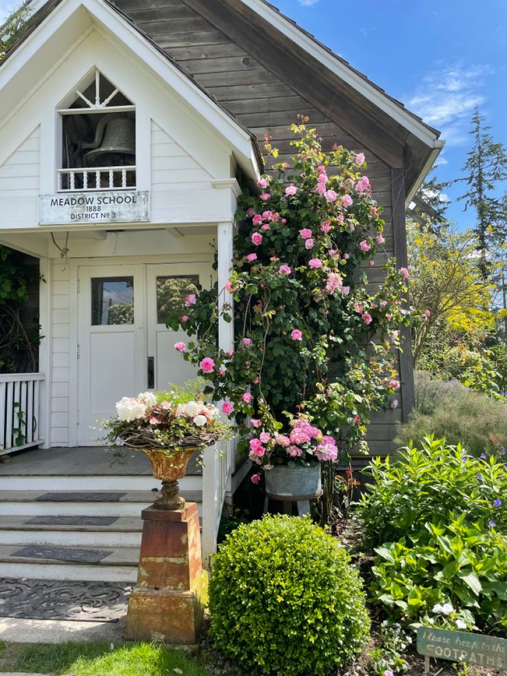 Historic schoolhouse with a blooming rose trellis and garden, white facade, bell tower, sunny day.