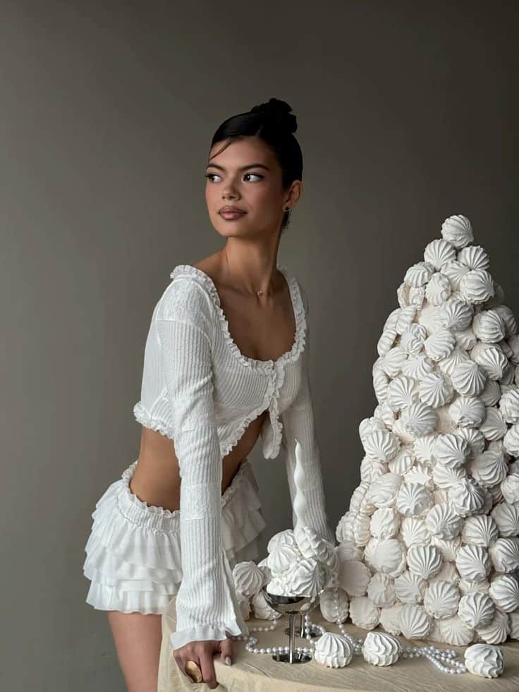 Woman in white outfit beside a meringue dessert tower on a table, stylish and elegant setting.