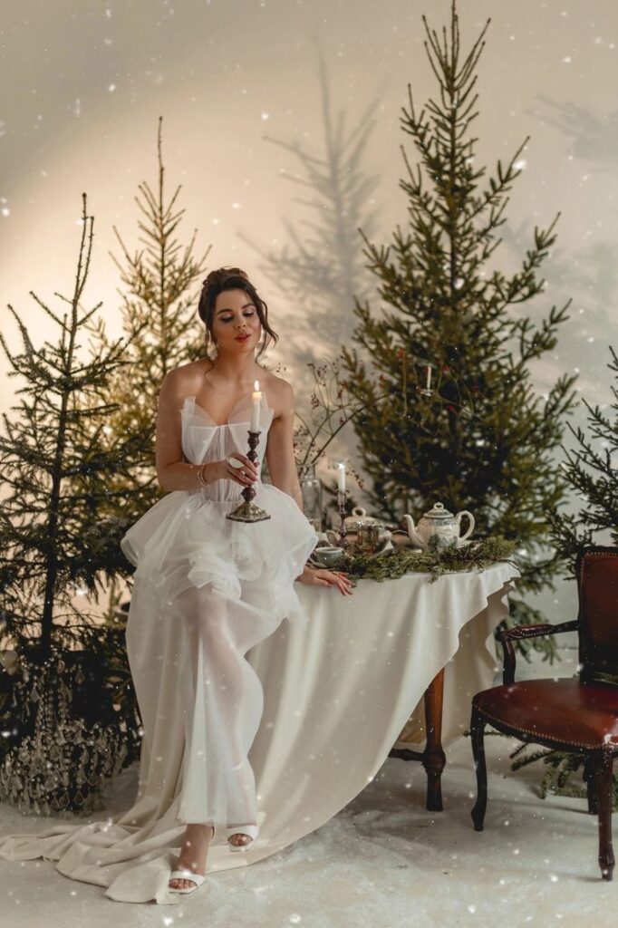 Elegant woman in white dress holding candle near festive table with pine trees in winter setting.