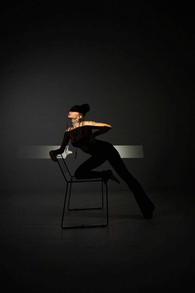 Silhouetted woman with tattoos leans on a chair in dramatic lighting, set against a dark background.