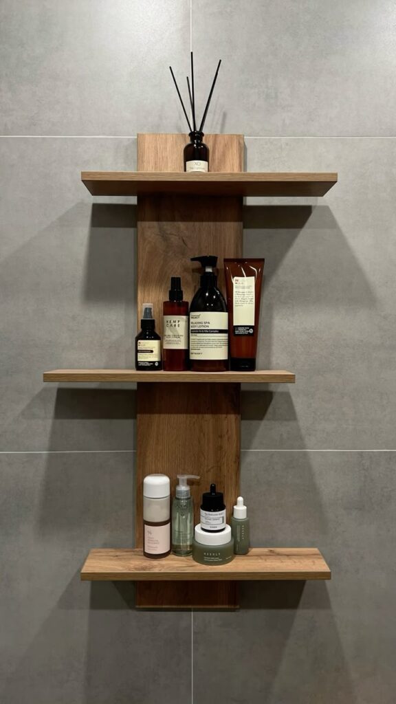 Wooden bathroom shelf with skincare products and aromatherapy diffuser on gray tile wall.