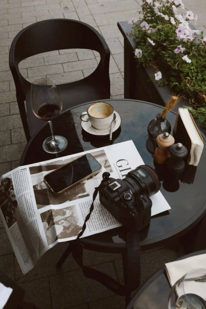 Outdoor café table with camera, coffee cup, wine glass, smartphone, and magazine.