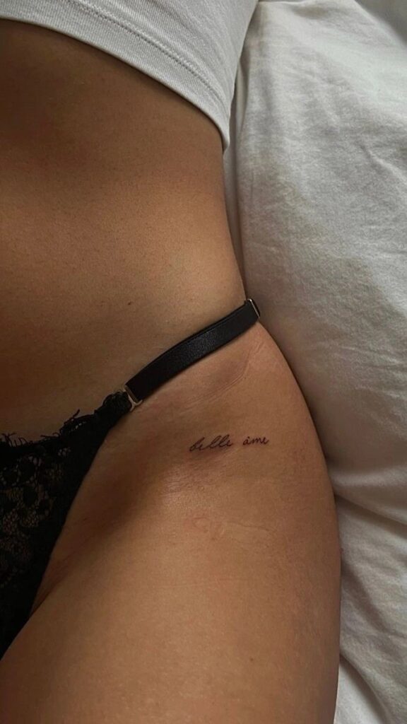 Small cursive tattoo on hip with lingerie strap, reading belle âme on skin next to white fabric.