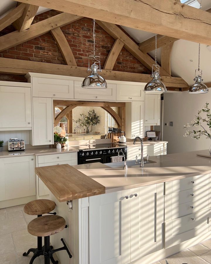 Rustic kitchen with wooden beams, cream cabinets, island, and vintage lighting in a sunlit space.