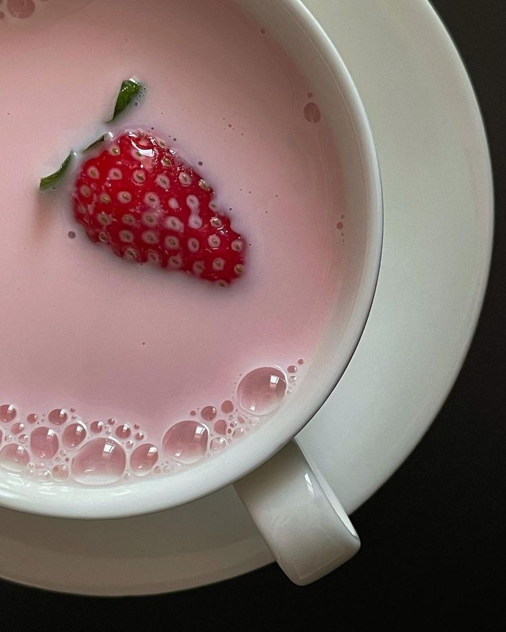 Strawberry floating in a cup of pink milk with bubbles, served on a white saucer.