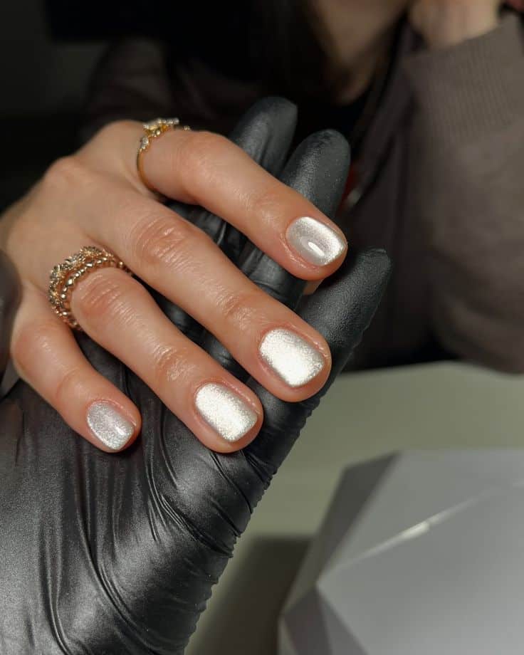Elegant white manicure with sparkly finish on short nails, held by a black gloved hand.