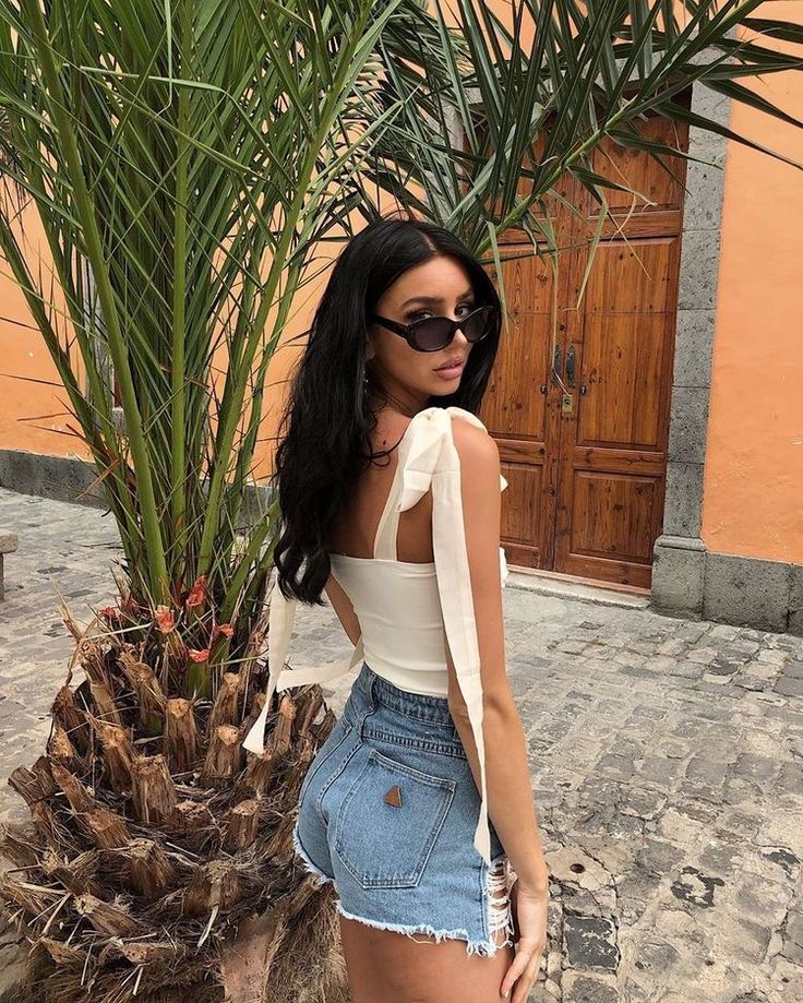 Woman in sunglasses and casual summer outfit standing near a palm tree with an orange wall in the background.