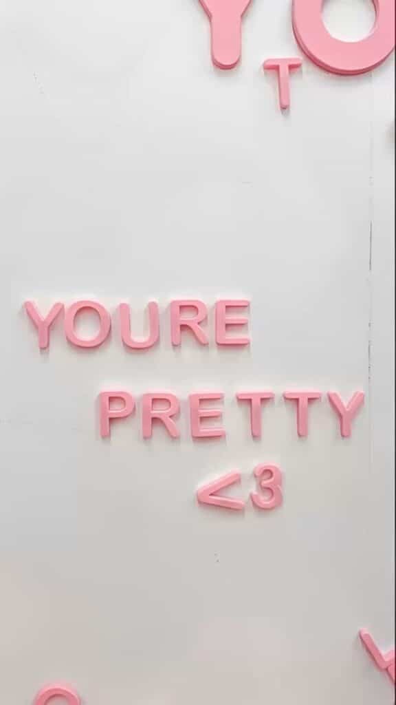 Pink letters spelling YOU'RE PRETTY <3 on a white wall.
