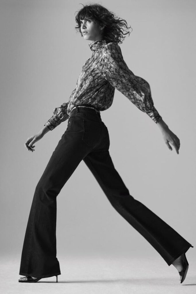 Fashionable woman in high-waisted jeans and patterned blouse walking confidently in heels, black and white photo.