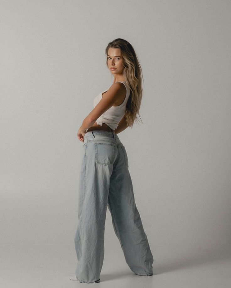 Woman modeling white tank top and wide-leg jeans in studio setting, minimalist background.