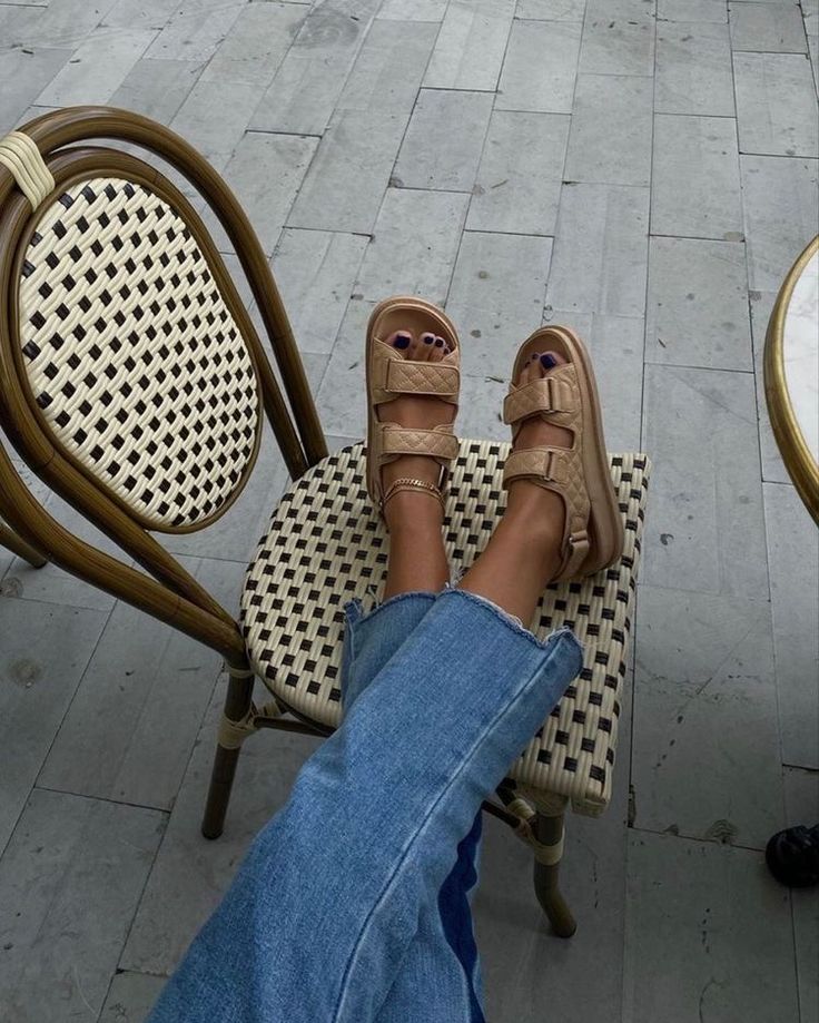 Relaxed feet in beige sandals rest on a woven chair, blue jeans casual style, outdoor setting.