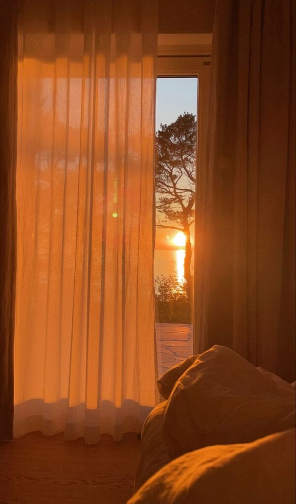 Cozy room with sheer curtains framing a stunning sunset view over trees and water. Warm, tranquil atmosphere.