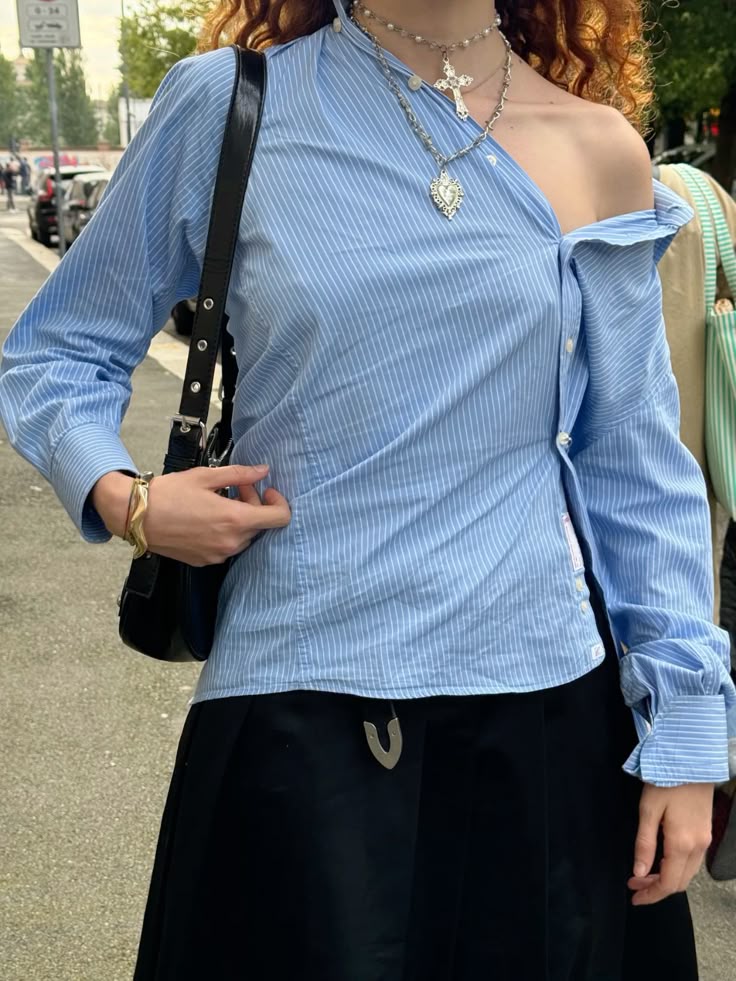Fashionable woman in blue pinstripe off-shoulder shirt and layered necklaces, holding a black bag on the street.