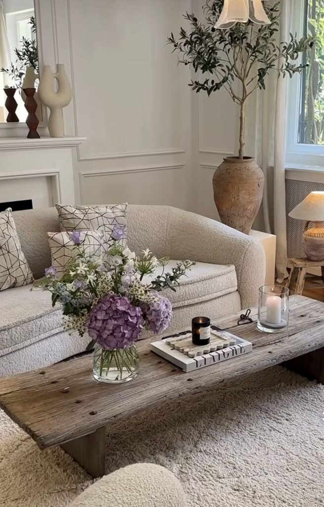 Stylish living room with cozy sofa, rustic table, candle, and purple flowers in an elegant vase for a serene ambiance.