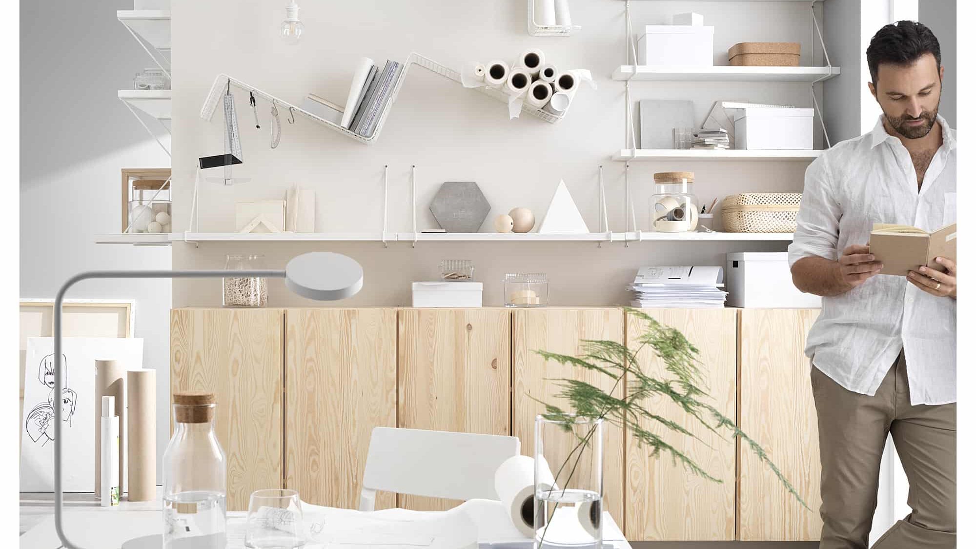 Modern home office with shelves, man reading. Minimalist design, natural wood cabinets, and desk decor.