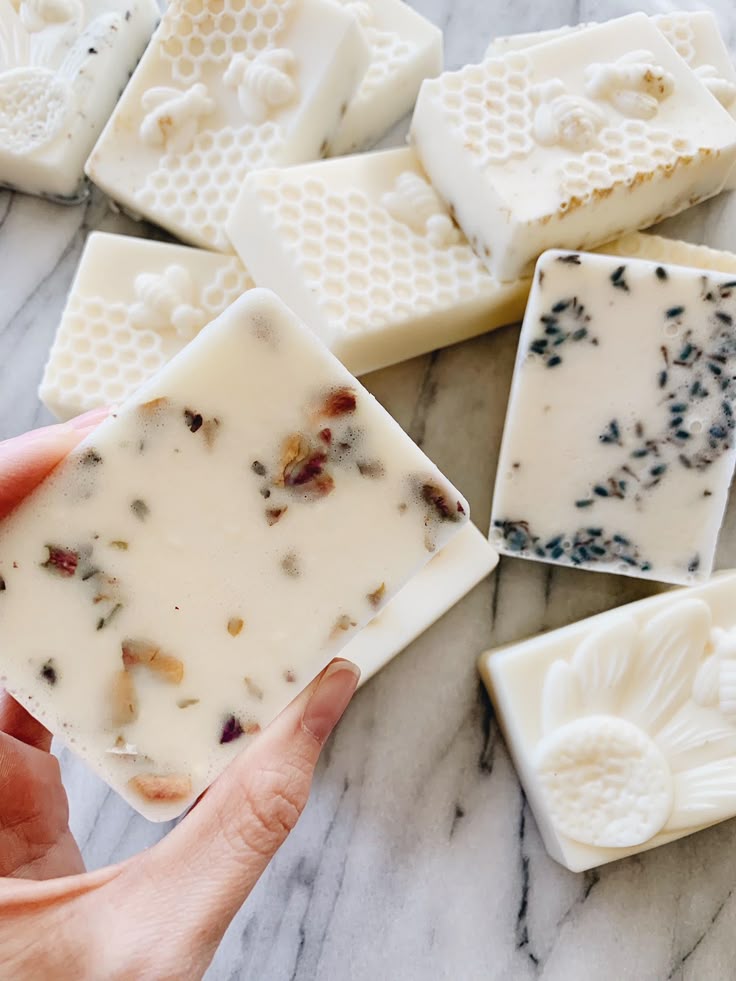 Hand holding a handcrafted soap bar with embedded natural ingredients, surrounded by various artisanal soaps on marble.