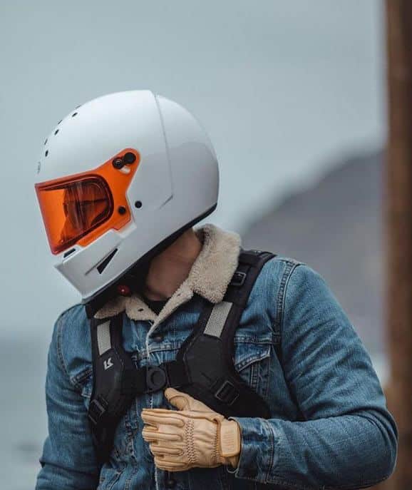 Motorcyclist in a white helmet and denim jacket, wearing gloves, with a scenic background.