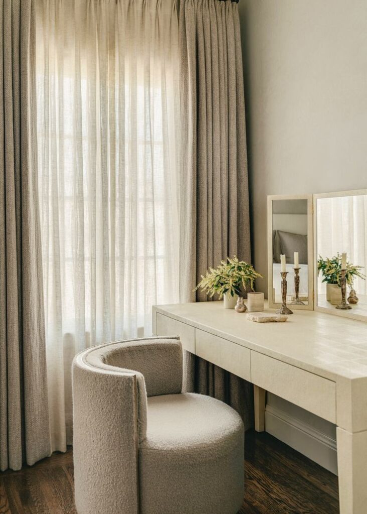 Cozy vanity corner with a beige chair, elegant curtains, and a wooden makeup table adorned with candles and plants.