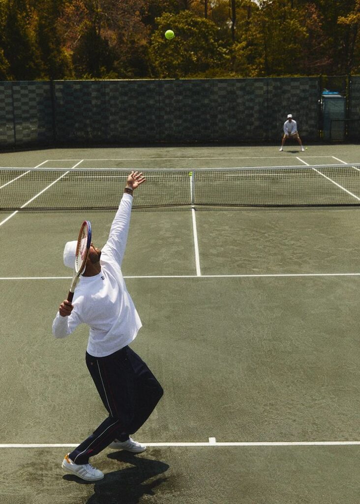 Player serving a tennis ball on an outdoor court, opponent ready to return, with trees in the background.