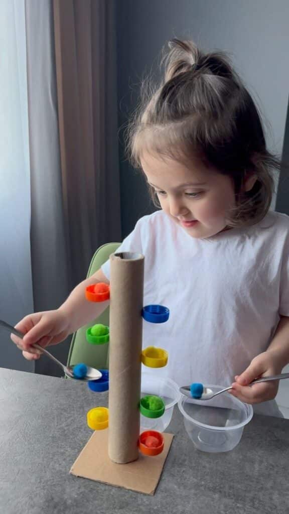 Child playing with homemade ball drop toy using spoons and colorful materials at a table.