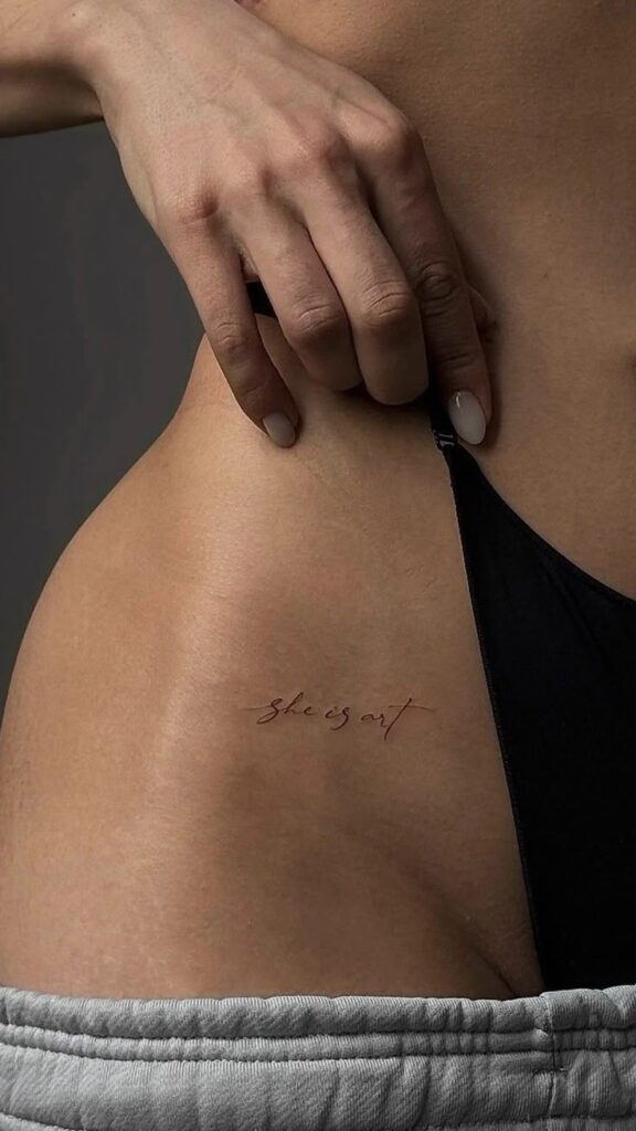 Close-up of a small tattoo above the hip that reads she is art, near the black strap of underwear.