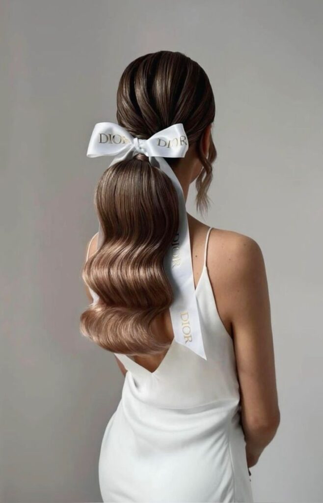 Elegant wavy ponytail tied with a branded white ribbon on a woman in a white dress, showcasing luxury style.