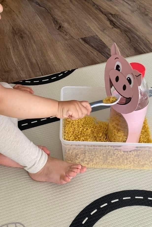 Child playing with pasta sensory bin, scooping macaroni into a pig-shaped container on a play mat.