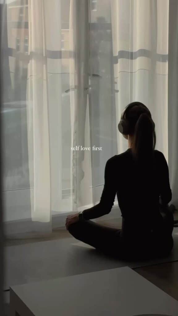 Person meditating by window with headphones, promoting self-love and calm reflection.
