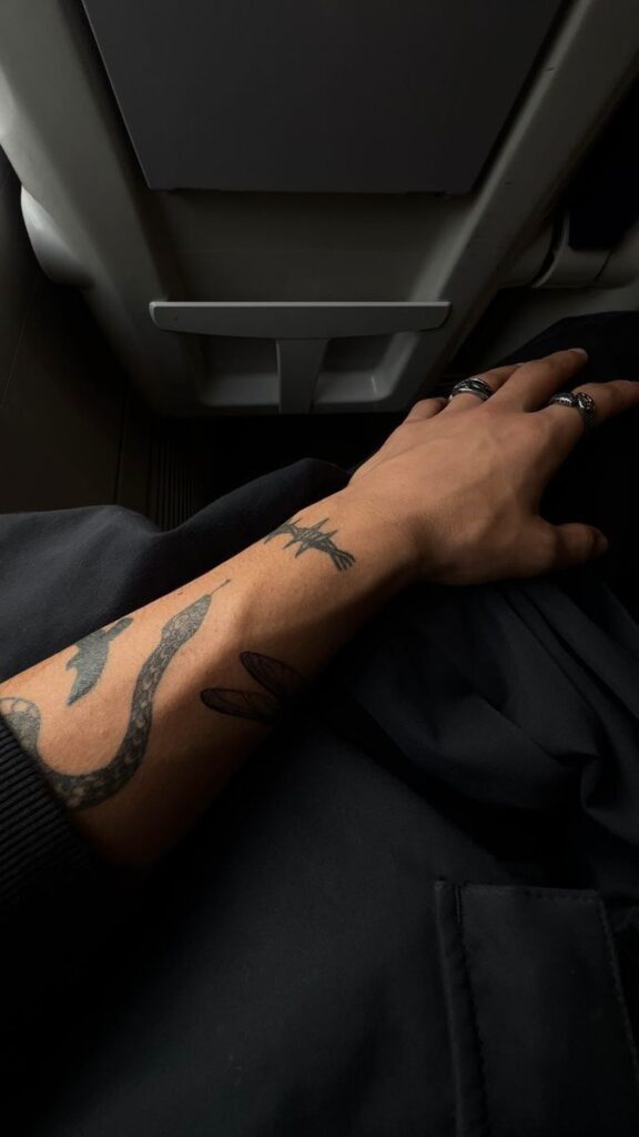 Tattooed arm with rings resting on dark fabric in airplane seat, highlighting unique designs and travel setting.