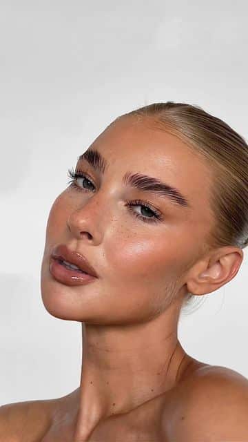 Close-up of a person with smooth skin, natural makeup, and styled hair, posing against a light background.