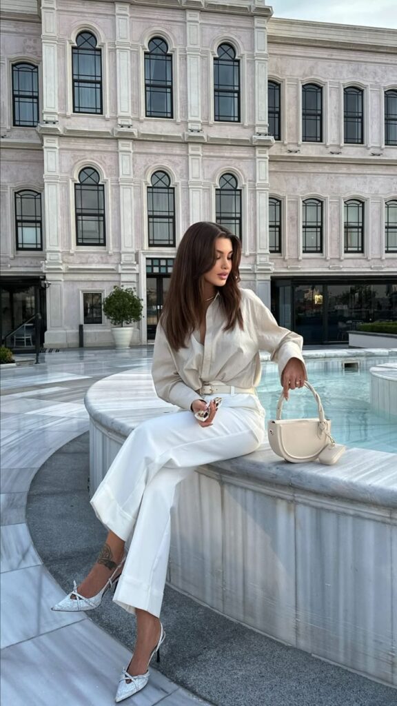 Woman in stylish outfit sitting by a fountain, holding a handbag, with an elegant building in the background.