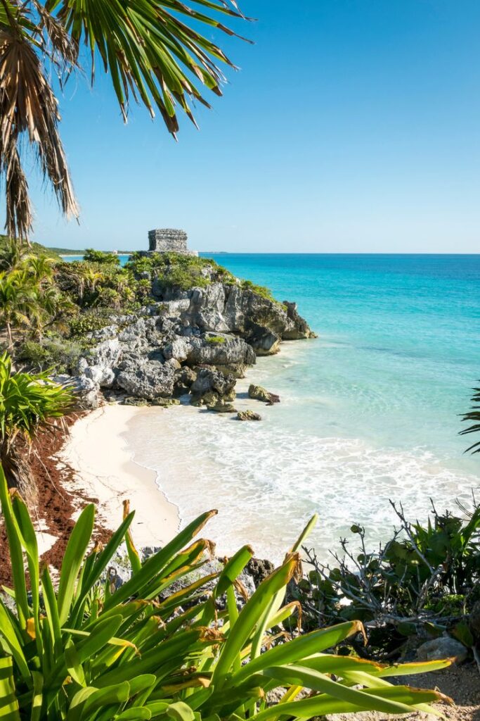 Tulum beach with turquoise water, palm trees, and ancient Mayan ruins under a clear blue sky.