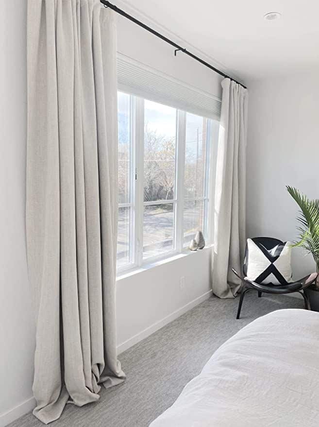 Bright bedroom with large window, beige curtains, white bedding, black chair, and plant, offering a cozy, modern look.