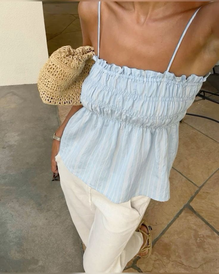Woman in light blue smocked top with white pants, holding a woven bag, standing on patio tiles. Fashion summer outfit.