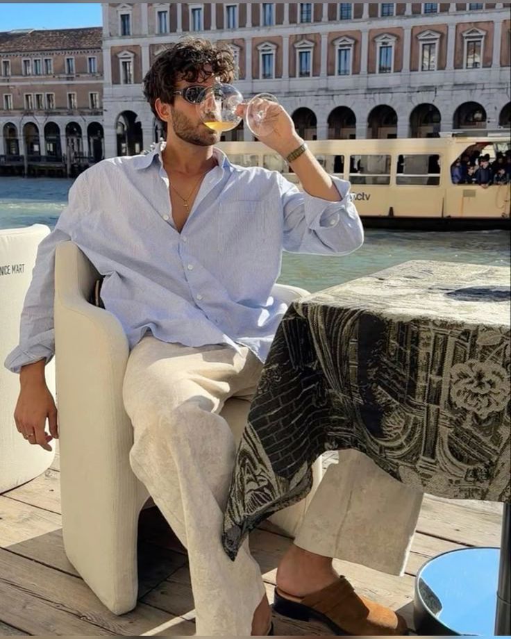 Man sipping wine by a canal in a stylish outfit, enjoying a sunny day near a historic European building.