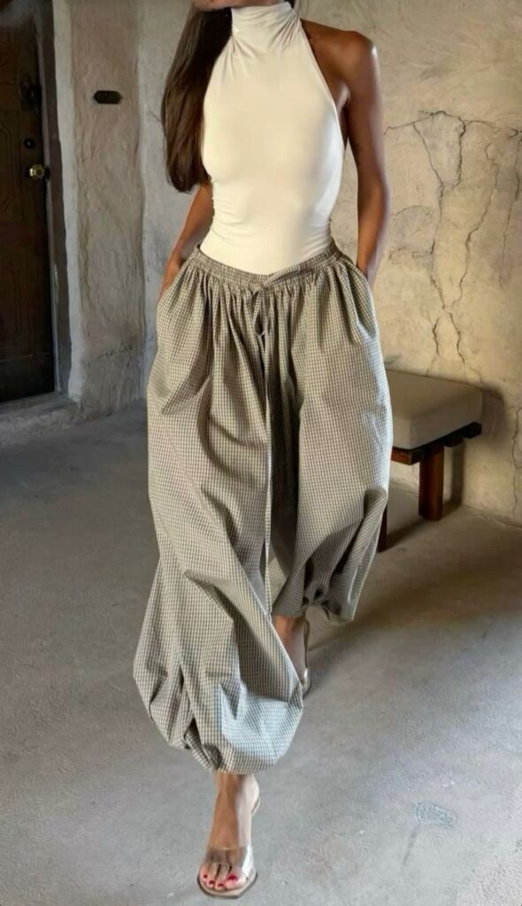 Woman in a sleeveless white top and plaid wide-leg pants, walking indoors on a rustic stone floor.