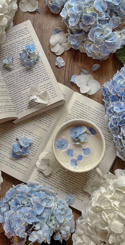 Open book with blue hydrangea petals and a tea cup, surrounded by blue and white flowers on a wooden table.