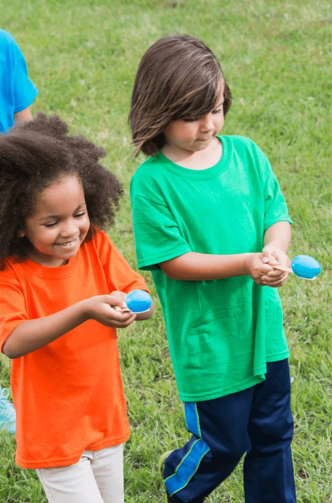 Children playing an egg-and-spoon race on green grass, smiling and wearing colorful shirts, showcasing teamwork and fun.