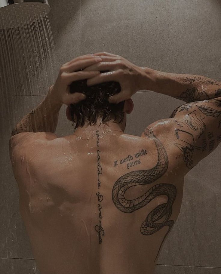 Man with tattoos showering, hands on head, displaying intricate snake design on back.